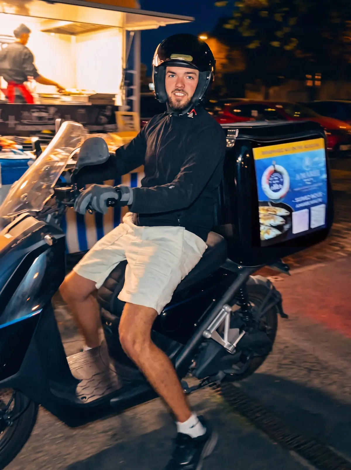 Le livreur du camion sur son scooter, le coffre noir aux couleurs d'A'Marée Minute, le camion éclairé derrière lui, de nuit.